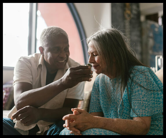 Elderly man gently helping woman drink while sitting together indoors.