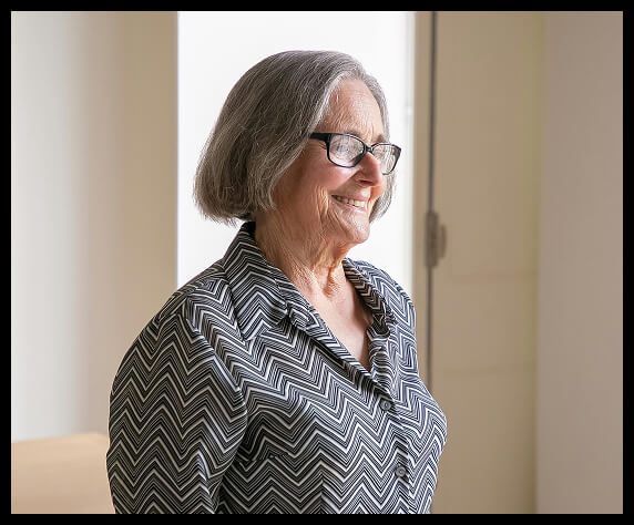 Elderly woman with silver hair, glasses, and a patterned blouse in a softly lit room.